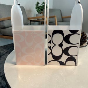 May include: Two upright rectangular storage bins with arched handles. One bin features a light pink and white repeating logo pattern, while the other displays a bold black and white repeating logo pattern. Both containers have white sides.
