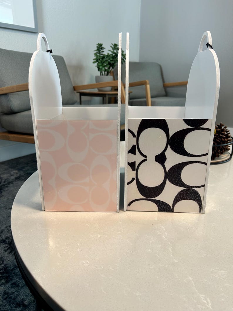 May include: Two upright rectangular storage bins with arched handles. One bin features a light pink and white repeating logo pattern, while the other displays a bold black and white repeating logo pattern. Both containers have white sides.