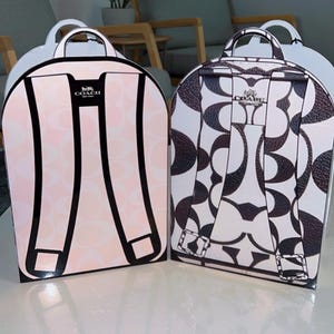 May include: Two Coach brand backpack-shaped gift boxes. The left box is light pink with a white signature C pattern and black strap illustrations. The right box is white with a dark brown signature C pattern and dark brown strap illustrations.