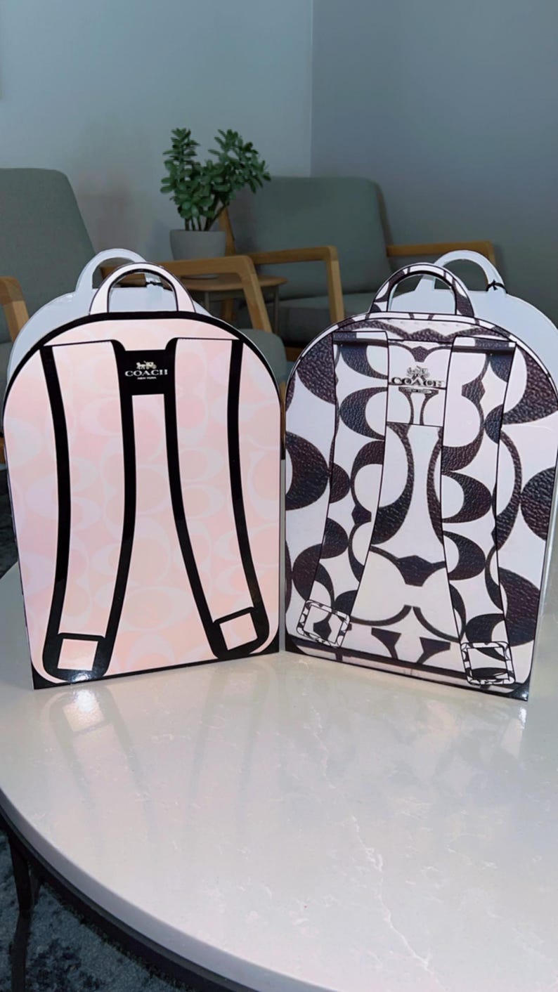 May include: Two Coach brand backpack-shaped gift boxes. The left box is light pink with a white signature C pattern and black strap illustrations. The right box is white with a dark brown signature C pattern and dark brown strap illustrations.