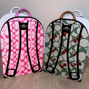 May include: Two flat, backpack-shaped cardboard display boxes. The left box features a bright pink Coach signature C pattern with a pink handle. The right box features a green Coach signature C pattern with red cherries and a tan handle.