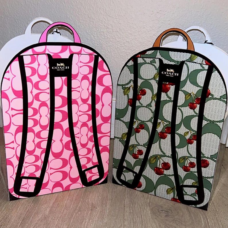 May include: Two flat, backpack-shaped cardboard display boxes. The left box features a bright pink Coach signature C pattern with a pink handle. The right box features a green Coach signature C pattern with red cherries and a tan handle.