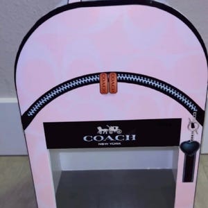 May include: A light pink backpack-shaped display box featuring a subtle tonal pattern. The front displays a black horizontal band with the white text COACH NEW YORK and a horse and carriage logo. It includes a black zipper detail and a heart charm.