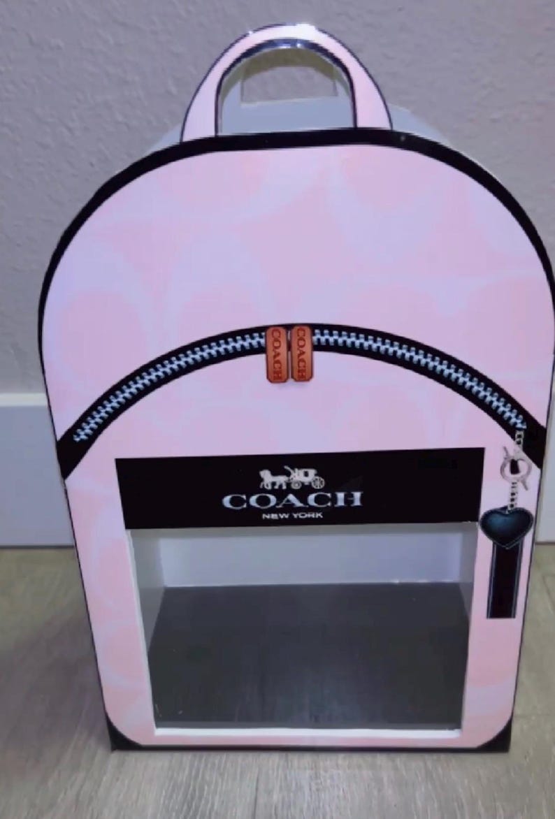 May include: A light pink backpack-shaped display box featuring a subtle tonal pattern. The front displays a black horizontal band with the white text COACH NEW YORK and a horse and carriage logo. It includes a black zipper detail and a heart charm.