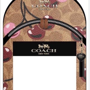 May include: A tan backpack featuring the Coach signature logo pattern with a pink cherry print. The bag has a top handle, a front zippered pocket with a black rectangular panel displaying the Coach New York logo, and a black heart-shaped charm.