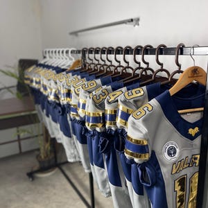 May include: A row of gray and navy blue football jerseys with gold accents hang on a rack. Each jersey has a number in gold and a team logo. The jerseys are displayed in a well-lit room.