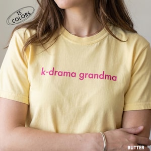 May include: A light yellow t-shirt with the text "k-drama grandma" in pink. The shirt is short-sleeved and has a round neckline. The words "15 COLORS" are in a white oval in the upper left corner, and the word "BUTTER" is in the lower right corner.