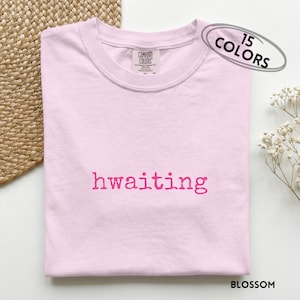 May include: A light pink t-shirt with the word "hwaiting" printed in a bright pink font. The shirt has a crew neck and a small tag at the neckline. The text "15 COLORS" is in an oval shape in the upper right corner.