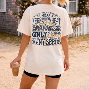 May include: A cream-coloured t-shirt with a graphic design. The text on the shirt reads: "VERY TRULY I TELL YOU UNLESS A KERNEL OF WHEAT FALLS TO THE GROUND AND DIES, IT REMAINS ONLY A SINGLE SEED (BUT IF IT DIES, IT PRODUCES MANY SEEDS) JOHN 12:24".