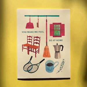 May include: Illustration featuring a broom, coffee pot, tennis rackets, and chairs. The text "YOU MAKE ME FEEL SO AT HOME" is written on the image. The color palette includes red, orange, green, and blue.