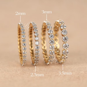 May include: Four gold eternity rings with clear gemstones. The rings are 2mm, 2.5mm, 3mm, and 3.5mm wide. The rings are displayed on a light brown surface.