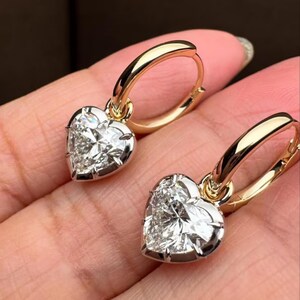 Könnte beinhalten: Ein Paar goldene Creolen mit herzförmigen Diamantanhängern. Die Diamanten sind in einem silberfarbenen Metall gefasst und funkeln hell. Die Ohrringe werden vor einem unscharfen Hintergrund gezeigt.