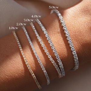 Op de afbeelding: Close-up van vijf tennisarmbanden met verschillende karaatgewichten: 1.0ctw, 2.0ctw, 3.0ctw, 4.0ctw en 5.0ctw. De armbanden zijn bezet met kleine, sprankelende stenen. Eén armband is goudkleurig, de andere zijn zilverkleurig.