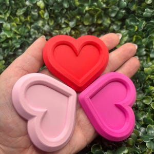 Könnte beinhalten: Drei herzförmige Ausstecher in Rot, Hellrosa und Pink. Die Ausstecher haben ein mehrschichtiges Design mit einem erhöhten Rand um die Herzform. Der Hintergrund ist ein Bett aus grünen Blättern.