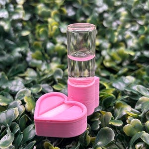 Puede incluir: Un vial de vidrio transparente con líquido rosa se encuentra sobre un recipiente rosa en forma de corazón con una franja blanca en el centro. Los objetos están colocados sobre un fondo de follaje verde.