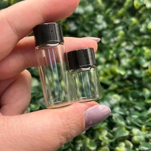 Peut inclure: Deux petits flacons en verre transparent avec des bouchons noirs. Les flacons sont pr&eacute;sent&eacute;s sur un fond de feuillage vert. L'un des flacons est l&eacute;g&egrave;rement plus grand que l'autre, et les deux sont vides.