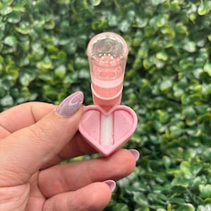 Peut inclure: Un applicateur rose en forme de c&oelig;ur avec un dessus cylindrique transparent. L'applicateur est tenu devant un fond de feuillage vert. La base en forme de c&oelig;ur a un insert blanc. L'applicateur est con&ccedil;u pour un usage cosm&eacute;tique.