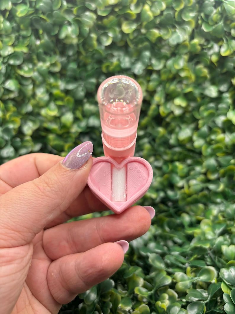 Peut inclure: Un applicateur rose en forme de c&oelig;ur avec un dessus cylindrique transparent. L'applicateur est tenu devant un fond de feuillage vert. La base en forme de c&oelig;ur a un insert blanc. L'applicateur est con&ccedil;u pour un usage cosm&eacute;tique.