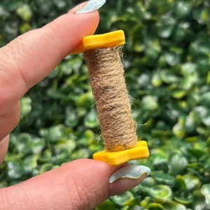 May include: A small, yellow spool holds a tightly wound roll of brown twine. The spool is held between two fingers against a backdrop of green foliage. The twine appears to be made of natural fibers.
