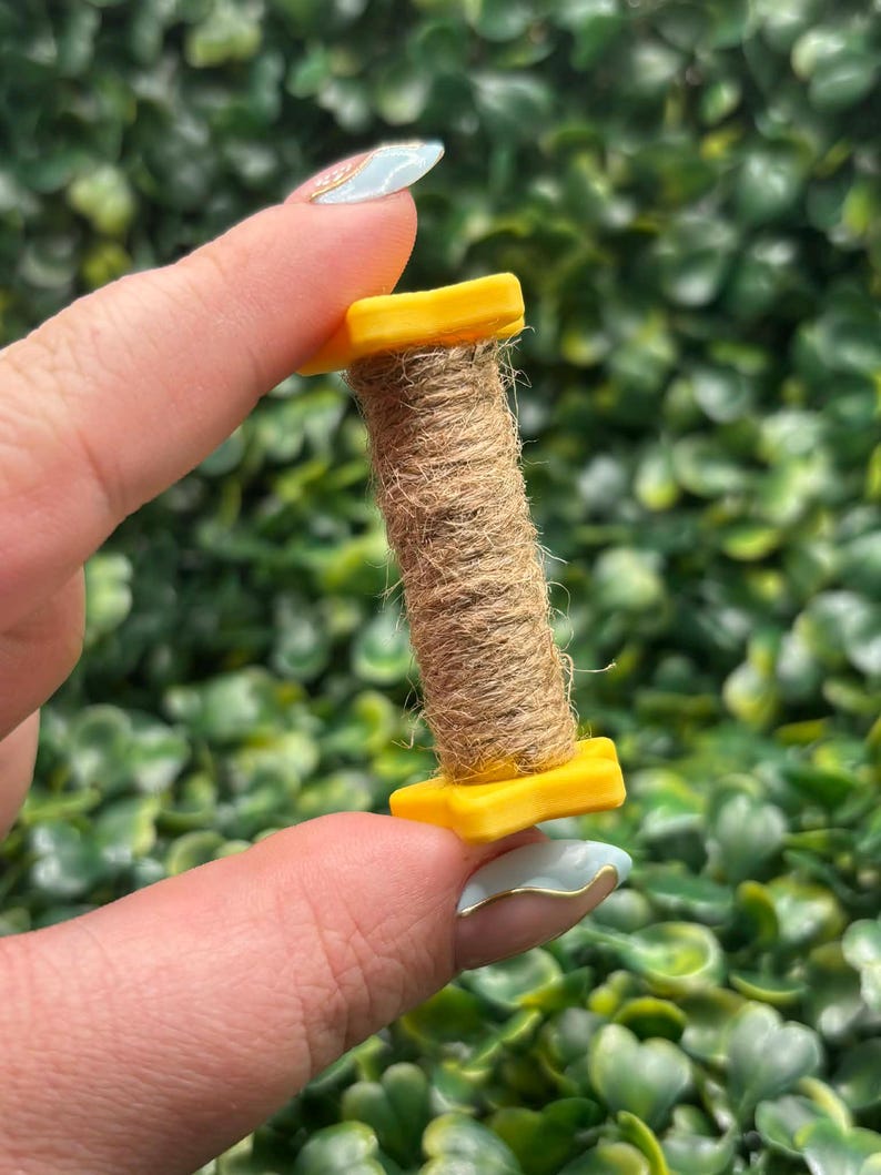 May include: A small, yellow spool holds a tightly wound roll of brown twine. The spool is held between two fingers against a backdrop of green foliage. The twine appears to be made of natural fibers.