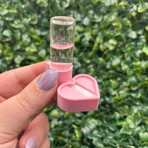 Peut inclure: Un petit flacon en verre transparent contenant un liquide rose est pos&eacute; sur une bo&icirc;te rose en forme de c&oelig;ur. La bo&icirc;te est ouverte, r&eacute;v&eacute;lant un objet rectangulaire blanc. Le fond est une haie verte dense.