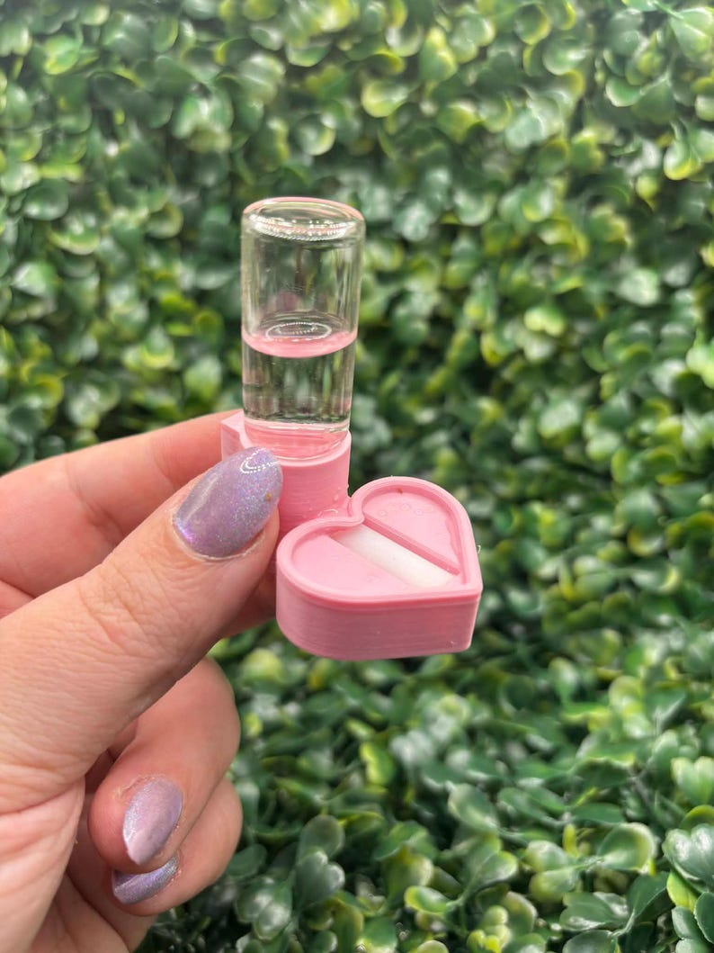 Peut inclure: Un petit flacon en verre transparent contenant un liquide rose est pos&eacute; sur une bo&icirc;te rose en forme de c&oelig;ur. La bo&icirc;te est ouverte, r&eacute;v&eacute;lant un objet rectangulaire blanc. Le fond est une haie verte dense.