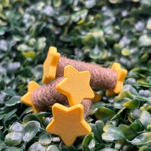 May include: Close-up of yellow star-shaped embellishments with a textured surface, attached to brown twine. The stars are scattered on a bed of green leaves, creating a natural and decorative aesthetic. Ideal for craft projects or adding a whimsical touch.