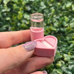 Peut inclure: Une petite bo&icirc;te rose en forme de c&oelig;ur avec un insert blanc, tenue &agrave; c&ocirc;t&eacute; d'un petit flacon transparent rempli de liquide rose. Les articles sont plac&eacute;s sur un fond de feuillage vert.