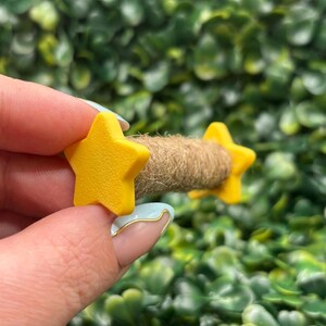 May include: A small, handmade craft item featuring two bright yellow star-shaped accents on either end of a brown, rope-like center. The stars have a smooth finish. The background is a dense, green, leafy texture.