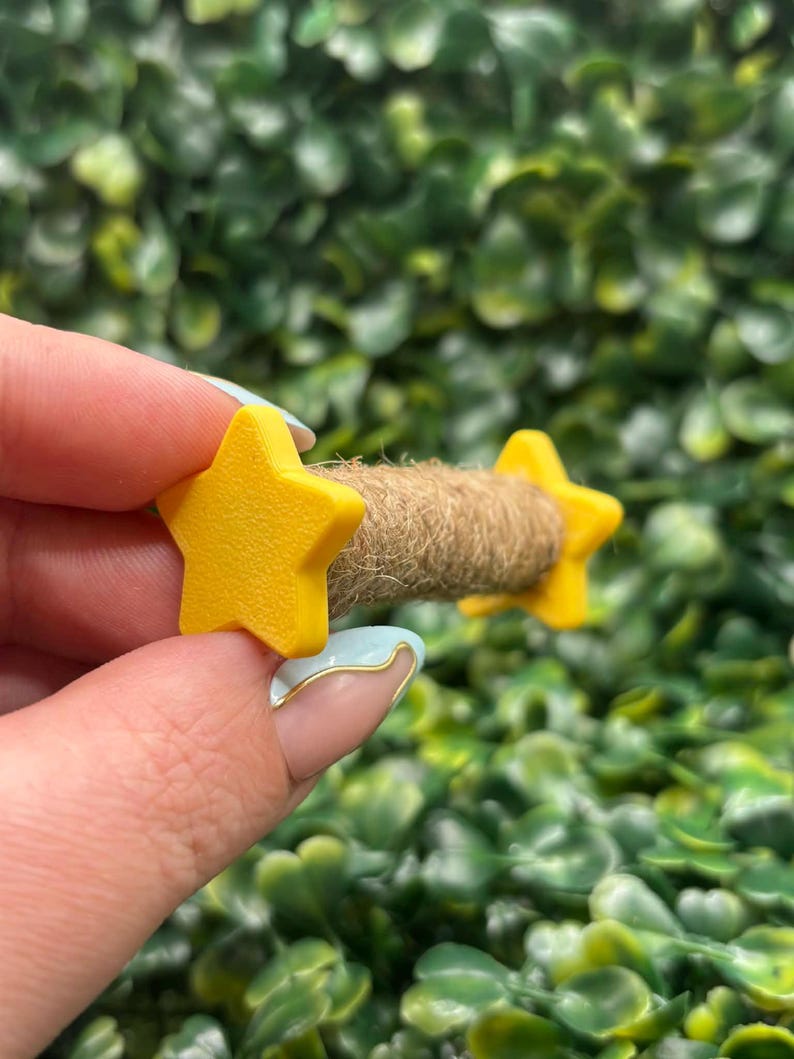 May include: A small, handmade craft item featuring two bright yellow star-shaped accents on either end of a brown, rope-like center. The stars have a smooth finish. The background is a dense, green, leafy texture.