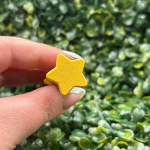 May include: A bright yellow star-shaped ring. The ring is held up against a backdrop of green foliage. The star has five points and a smooth surface. The ring is a simple, playful accessory.