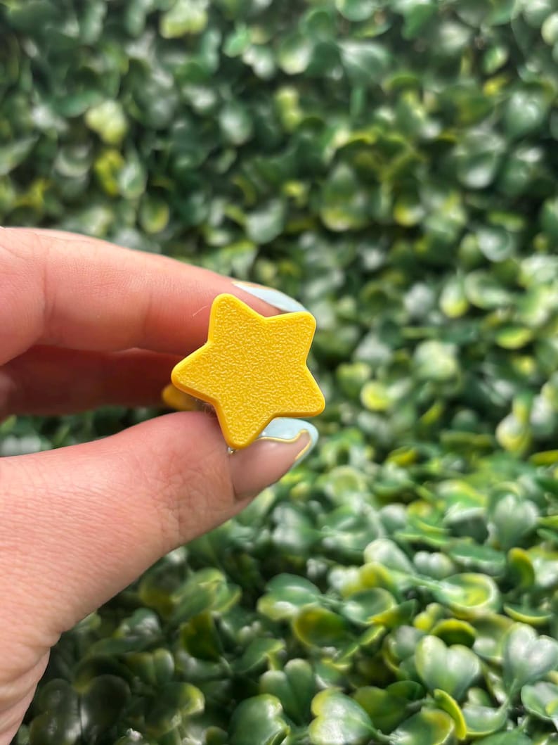 May include: A bright yellow star-shaped ring. The ring is held up against a backdrop of green foliage. The star has five points and a smooth surface. The ring is a simple, playful accessory.