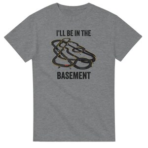 May include: A heather gray t-shirt with the text "I'LL BE IN THE BASEMENT". The shirt features a graphic of a black and yellow race track design.