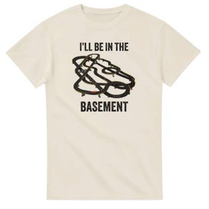 May include: Cream-colored t-shirt with the text "I'll be in the basement" in black block letters. A black and gold abstract design is centered above the word "basement". The shirt has short sleeves and a crew neck.