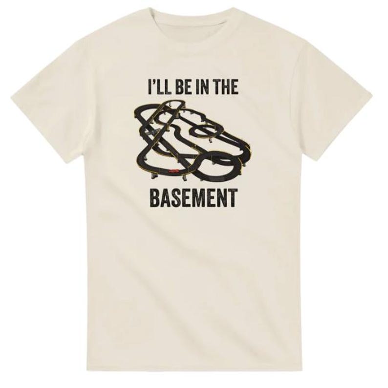 May include: Cream-colored t-shirt with the text "I'll be in the basement" in black block letters. A black and gold abstract design is centered above the word "basement". The shirt has short sleeves and a crew neck.