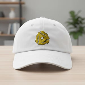 May include: A white baseball cap with a gold-colored 45 RPM record adapter embroidered on the front. The hat has a curved brim and a classic six-panel design. The cap is made of a soft, durable material.