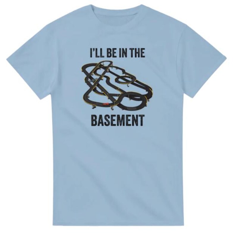 May include: Light blue t-shirt with the text "I'LL BE IN THE BASEMENT" above a graphic of a black and gold race track. The shirt is made of a soft material and has short sleeves.
