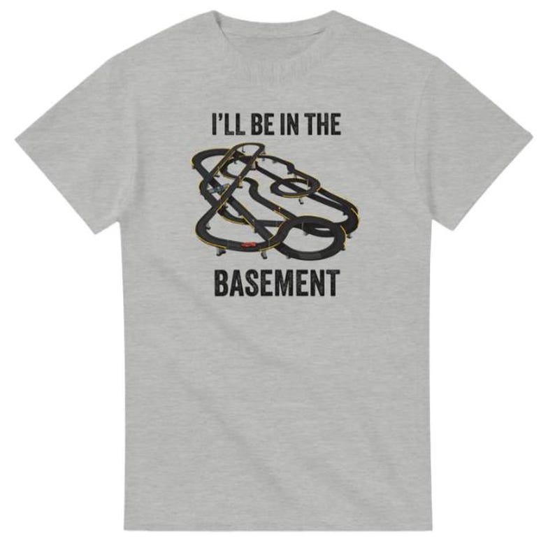 May include: A light gray t-shirt with the text "I'LL BE IN THE BASEMENT" in black. The shirt features a graphic of a black and gold slot car racing track.