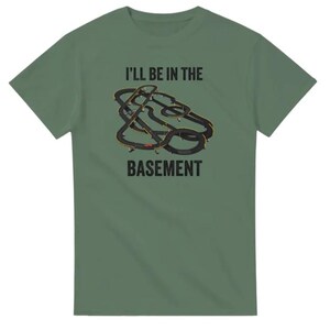 May include: Green t-shirt with the text "I'LL BE IN THE BASEMENT" above a graphic of a slot car race track. The track is black with yellow and red accents. The shirt is a solid green color.