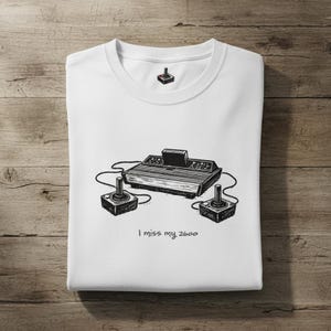 May include: A white t-shirt featuring a black ink illustration of a vintage video game console with two joysticks and a cartridge. The text "I miss my 2600" is printed below the console, and a small joystick icon is located at the inner neckline.