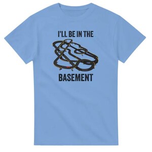 May include: Light blue t-shirt with the text "I'll be in the basement" above a graphic of a slot car race track. The track is black with yellow and red accents. The shirt is a short-sleeved crew neck.