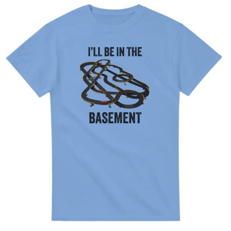 May include: Light blue t-shirt with the text "I'll be in the basement" above a graphic of a slot car race track. The track is black with yellow and red accents. The shirt is a short-sleeved crew neck.