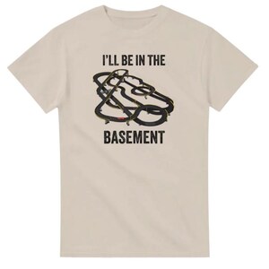 May include: Beige t-shirt with the text "I'LL BE IN THE BASEMENT" above a graphic of a black and yellow race track. The t-shirt is made of a soft, comfortable material.