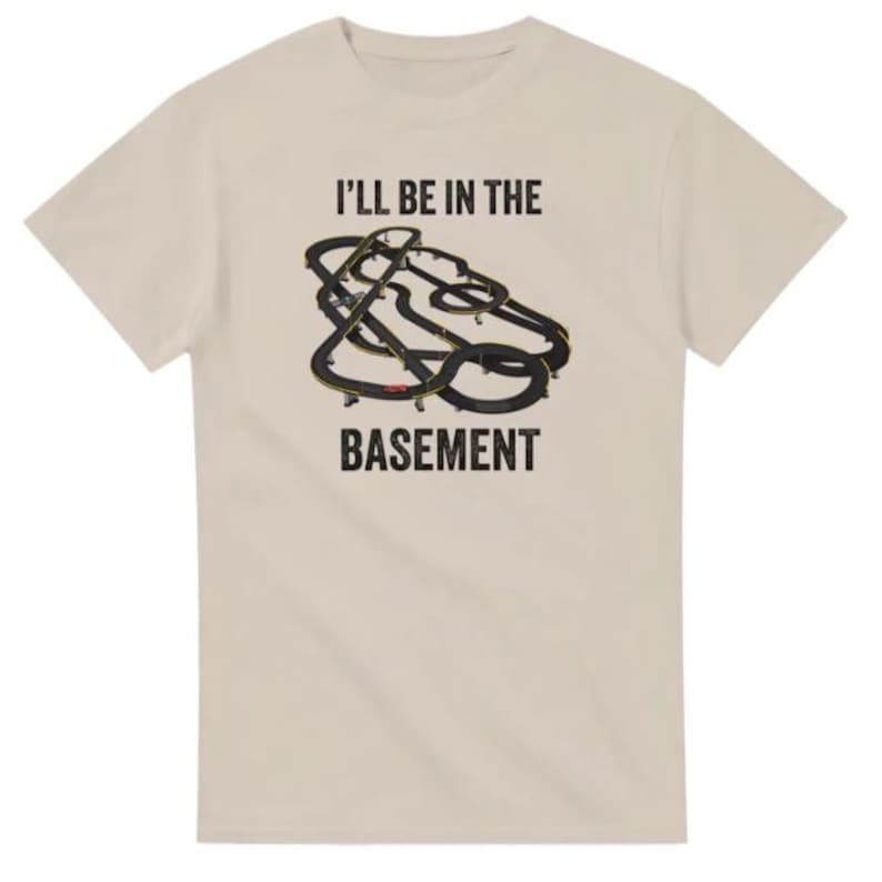 May include: Beige t-shirt with the text "I'LL BE IN THE BASEMENT" above a graphic of a black and yellow race track. The t-shirt is made of a soft, comfortable material.