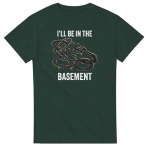May include: Dark green t-shirt with the text "I'll be in the basement" in white. The shirt features a graphic of a race track in black and gold.