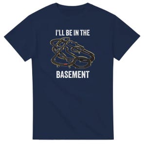 May include: Navy blue t-shirt with the text "I'll be in the basement" above a graphic of a race track. The graphic is black and gold. The t-shirt is a short-sleeved crew neck.