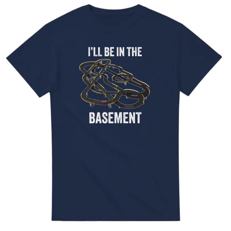 May include: Navy blue t-shirt with the text "I'll be in the basement" above a graphic of a race track. The graphic is black and gold. The t-shirt is a short-sleeved crew neck.