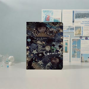 May include: A rectangular notebook featuring a dark blue collage cover with Coraline characters, a black cat, a planet, a clock, a camera, and stars. Text includes "Be wise. Be brave. Be tricky.", "Is this a memory or a dream?", and "Coraline".