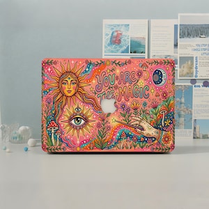 Può includere: Una skin per laptop con uno sfondo rosa vivace e un'illustrazione colorata di un sole, una luna crescente, un occhio, funghi, fiori e una mano che tiene una bacchetta. Il testo "YOU ARE THE MAGIC" è stampato in un carattere stilizzato al centro.