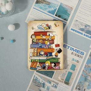 May include: A rectangular decorative sign featuring a colorful illustration of Peanuts characters interacting with a large stack of books. The scene includes Snoopy, Charlie Brown, Lucy, Linus, and Woodstock, with a vintage-style aged paper background.